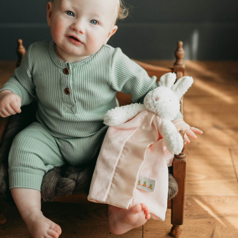 Baby in green outfit holding a pink bunny security blanket for babies