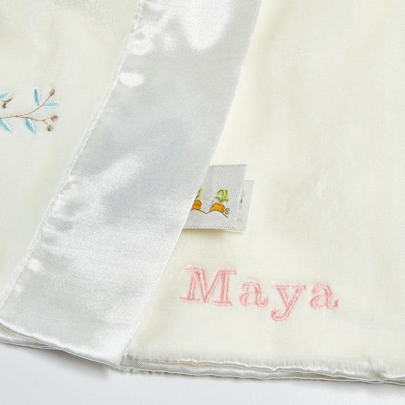 Close up view of buddy blanket custom embroidery with the name 'Maya' embroidery in pink on a white background