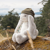 A plush white bunny toy wearing an olive green knit beanie sits on a mossy rock outdoors with blurred trees and sky in the background.