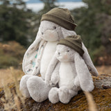 Two plush bunny toys, one larger and one smaller, sitting together on a mossy rock outdoors. The bunnies are light gray and white with olive green knit beanies, set against a blurred natural background of trees and grass.