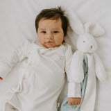 Baby in a white onesie lying next to a plush bunny buddy blanket toy on a soft surface