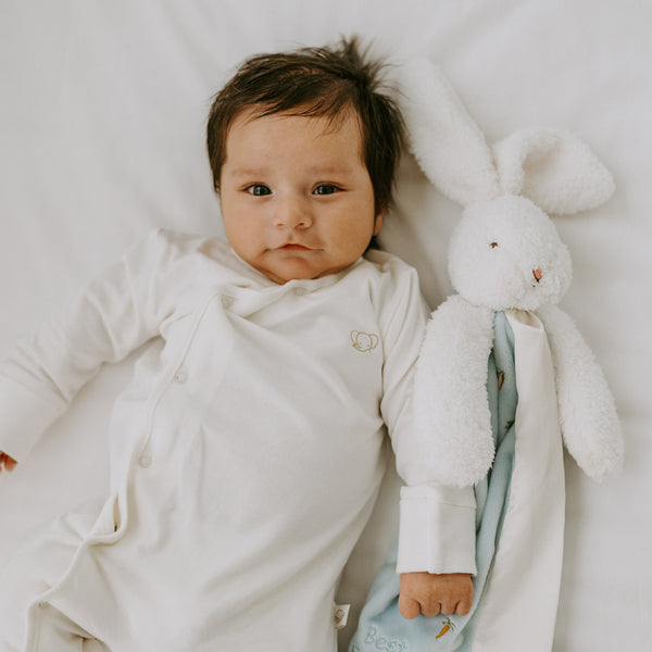 Baby in a white onesie lying next to a plush bunny toy on a soft surface