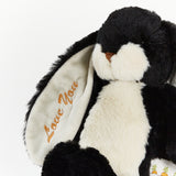 A close-up of a plush bunny toy with black and white fur, a small brown stitched nose, and orange embroidered text on the inner ear reading 'Love You'.