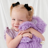 A smiling toddler with blonde pigtails and black bow hair clips hugs a large, fluffy lavender purple plush toy against a soft white curtain background.
