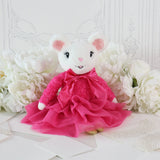 A white plush mouse toy wearing a bright pink ruffled dress and bow, posed among large white flowers on a light surface.
