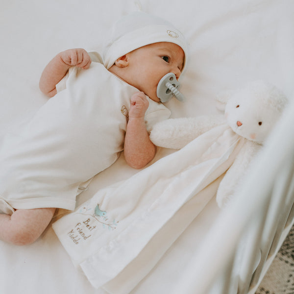 Newborn baby wrapped in a white blanket with a pacifier, lying on a soft surface.