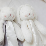 Lifestyle image of Two white plush bunny toys, attached blanket body with pink cheeks styled on a white background