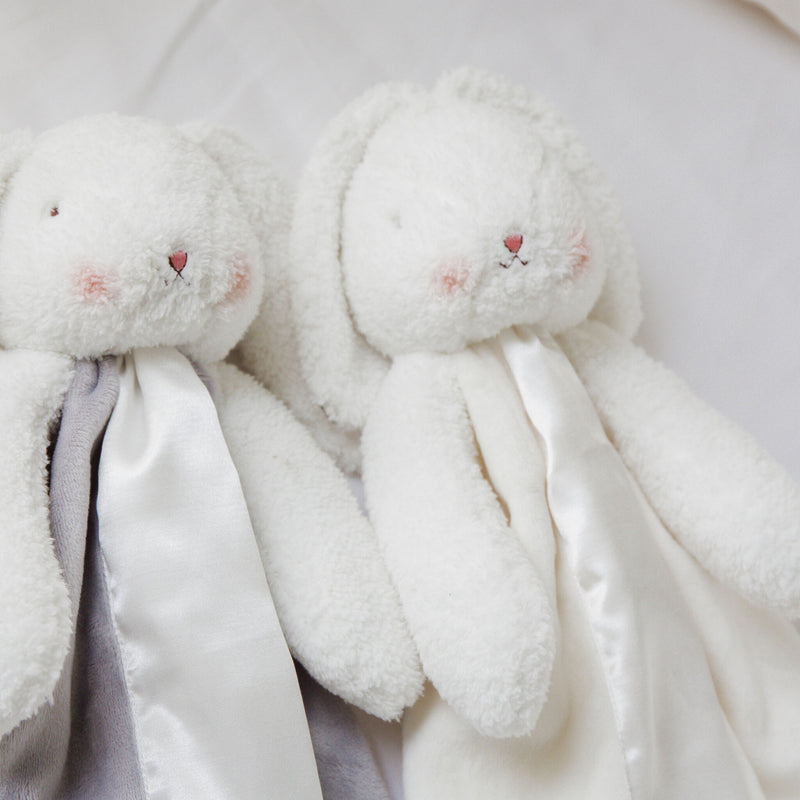 Lifestyle image of Two white plush bunny toys, attached blanket body with pink cheeks styled on a white background