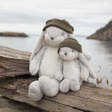 Two plush bunny toys in soft white plush fabric, each wearing an olive green knit hat, sitting on a weathered log with a blurred coastal water and land background.