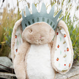 A plush stuffed bunny with long ears and a crown, photographed outdoors against soft greenery and flowers. The bunny is cream and tan with white inner ears and small colorful printed motifs inside the ears.