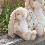 A beige plush bunny stuffed animal sitting on a weathered wooden surface outdoors, with tall green grass in the background. A second stuffed bunny is partially visible behind it. Both bunnies have patterned ears.