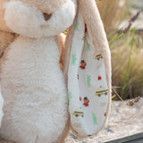 Close-up of a plush bunny toy with a cream-colored fabric ear flap printed with small colorful illustrations of taxis, apples, bears and the Statue of Liberty, set against a blurred outdoor background.