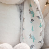 Close-up of a plush stuffed animal with soft cream fur and a patterned inner ear. The ear features a light cream background with small printed woodland motifs in mint green, gray, and dark gray.