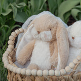 Two plush bunnies in a woven basket with wooden beads against a green leafy background