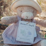 Taupe bunny plush in a purple dress holding a 'Bunnies by the Bay Seedlings' tag against a natural background.