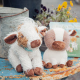 Two plush toys resembling cows on a rustic surface with flowers in the background.