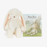A cream-colored plush bunny toy with long floppy ears sits beside a children's book titled 'Bun Bun: A Long Story' on a clean white background.
