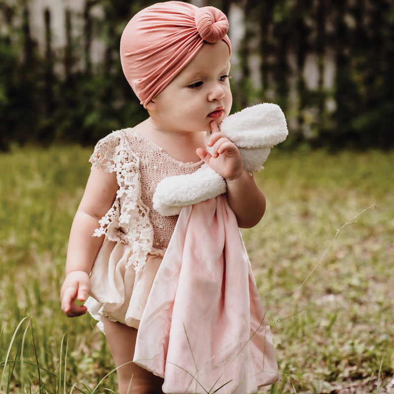 Baby in a pink outfit with a headband standing in a grassy field.