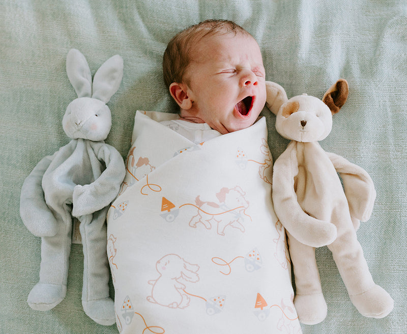 Swaddled baby yawning in bed with stuffed animals